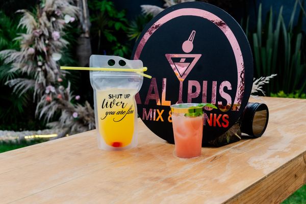 Cóctel en bolsa con popote y cóctel en vaso, frente al letrero de ALYPUS