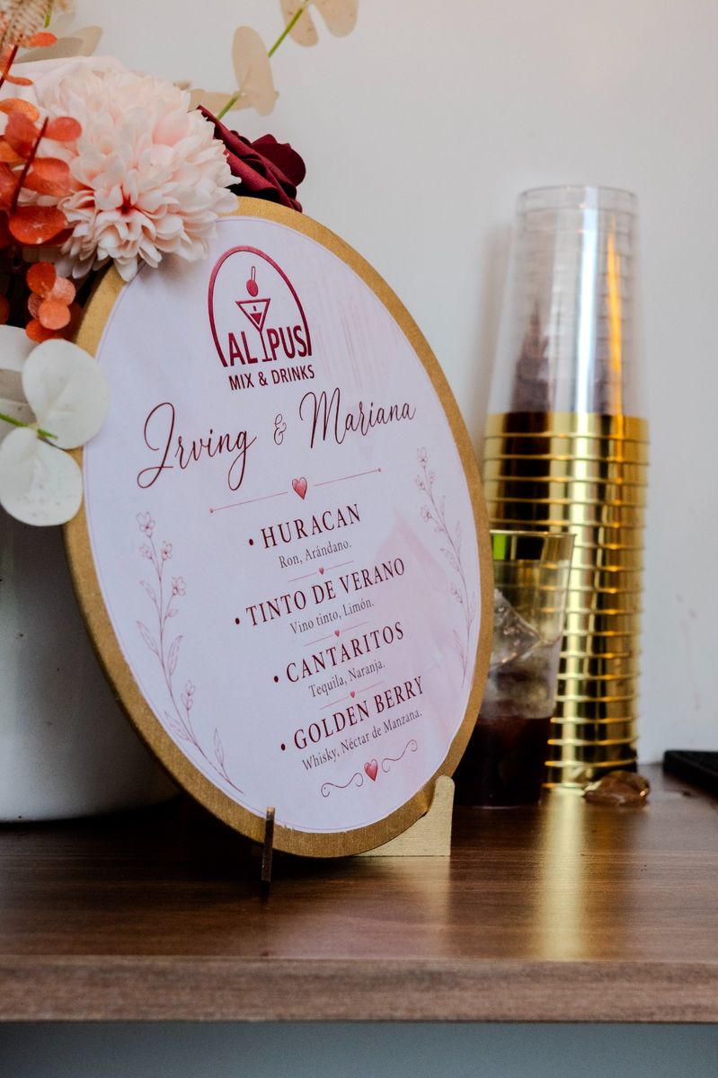 Letrero con la carta de cócteles de una boda, junto a flores y vasos de borde dorado